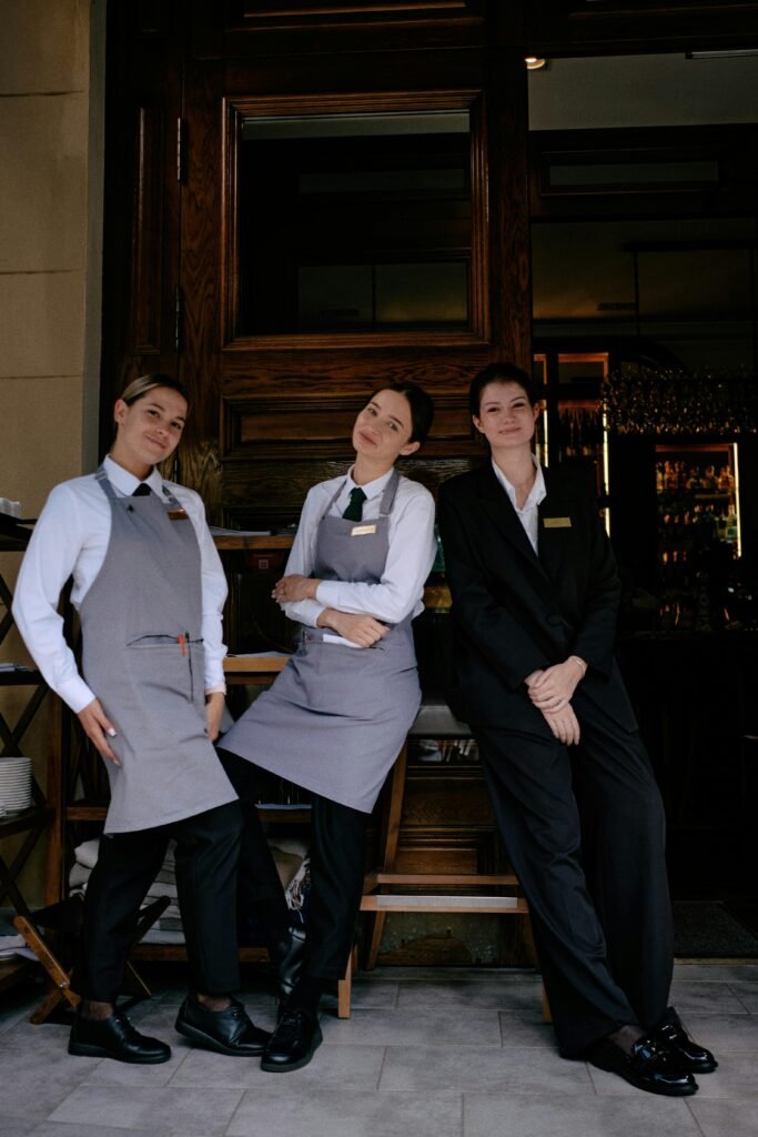 Three restaurant staff members in formal uniforms posing cheerfully in front of a restaurant.