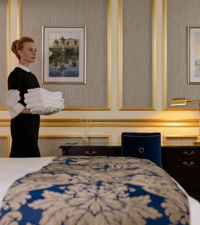 A housekeeper holding towels in a luxurious hotel room with elegant decor and a classic desk.