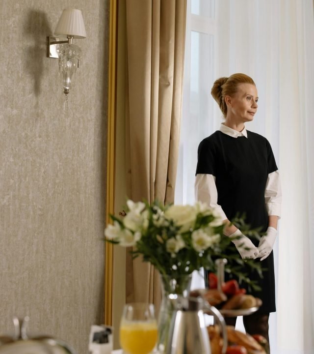 Elegantly dressed staff stands ready in a hotel room with a breakfast tray.